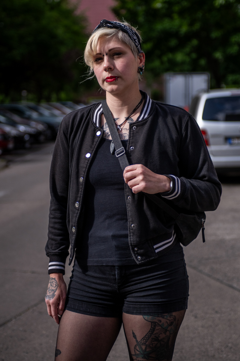 Die Aufnahme zeigt eine junge Frau, vermutlich im Alter von 25-35 Jahren, die in einer belebten Straße steht. Ihre Kleidung ist vollständig schwarz – eine schwarze Baseballjacke mit weißen Details, ein schwarzes T-Shirt, schwarze Shorts und schwarze Strumpfhosen. Ihre roten Lippen und das Bandana im Haar verleihen ihr einen rebellischen Look. Sie trägt eine schwarze Schultertasche über der Schulter. Im Hintergrund ist ein grauer Kleinwagen (wahrscheinlich ein VW Golf oder ähnliches) und ein typisches europäisches Reihenhaus zu sehen. Die Szene wirkt urban und modern.