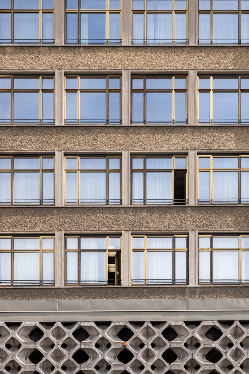 Die Aufnahme zeigt eine detailreiche Perspektive auf ein mehrgeschossiges Wohngebäude. Die Fassade ist in einem gedämpften Braunton gehalten, was dem Bild eine düstere, fast monochrome Atmosphäre verleiht. Die Fenster sind groß und rechteckig, mit dunklen Holzrahmen. Die Fassade selbst weist eine filigrane, geometrische Struktur auf, die aus Beton gefertigt ist. Im Vordergrund ist ein einzelnes Fenster geöffnet, wodurch ein Lichtstreifen in den Himmel hinein fällt. Die Szene wirkt ruhig und kontemplativ.