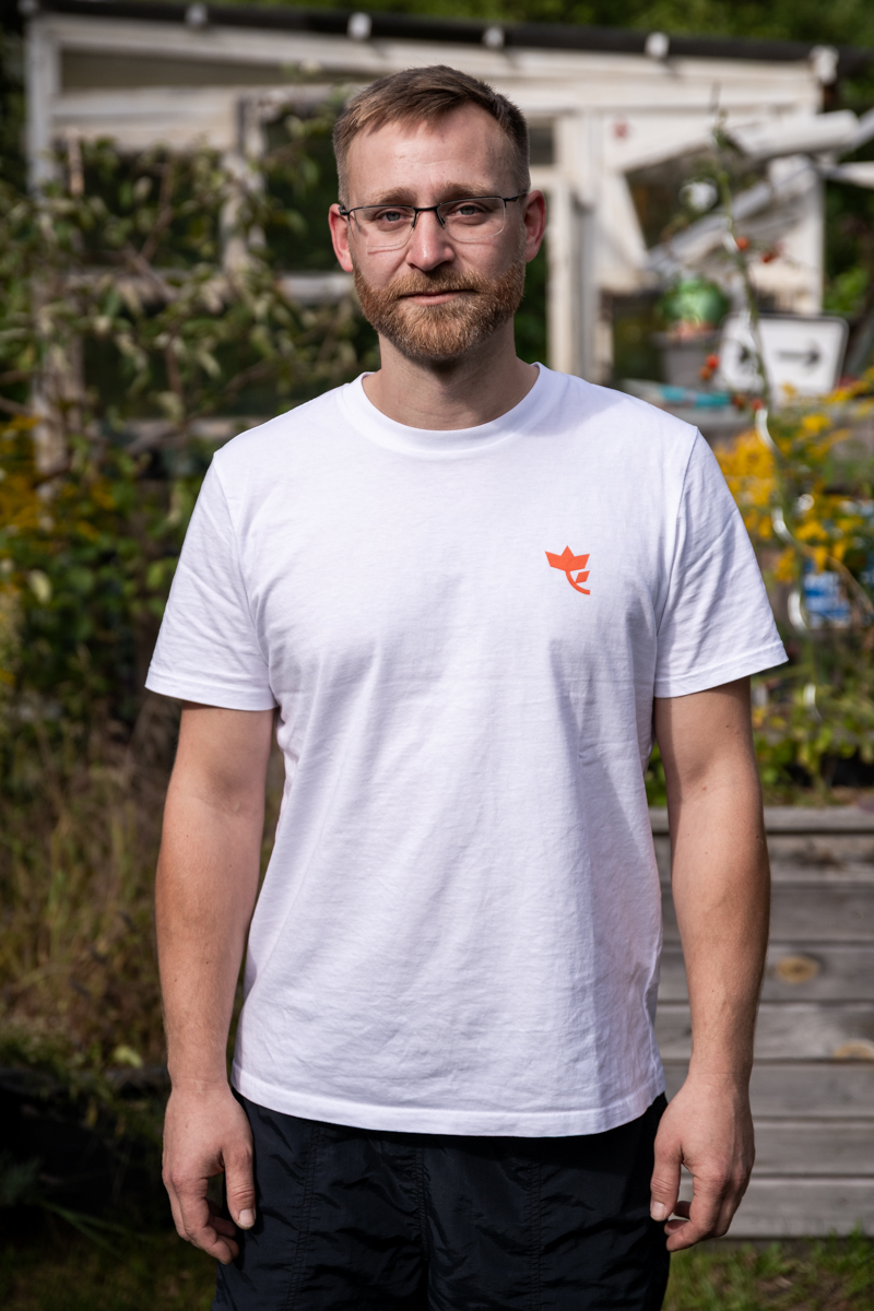 Die Aufnahme zeigt einen Mann, der vor einem Gewächshaus steht. Er trägt ein weißes T-Shirt mit einem roten Logo auf der Brust. Er trägt eine Brille und eine schwarze Hose. Im Hintergrund befindet sich ein Gewächshaus mit einem Metallgitter. Die Szene wirkt entspannt und natürlich.