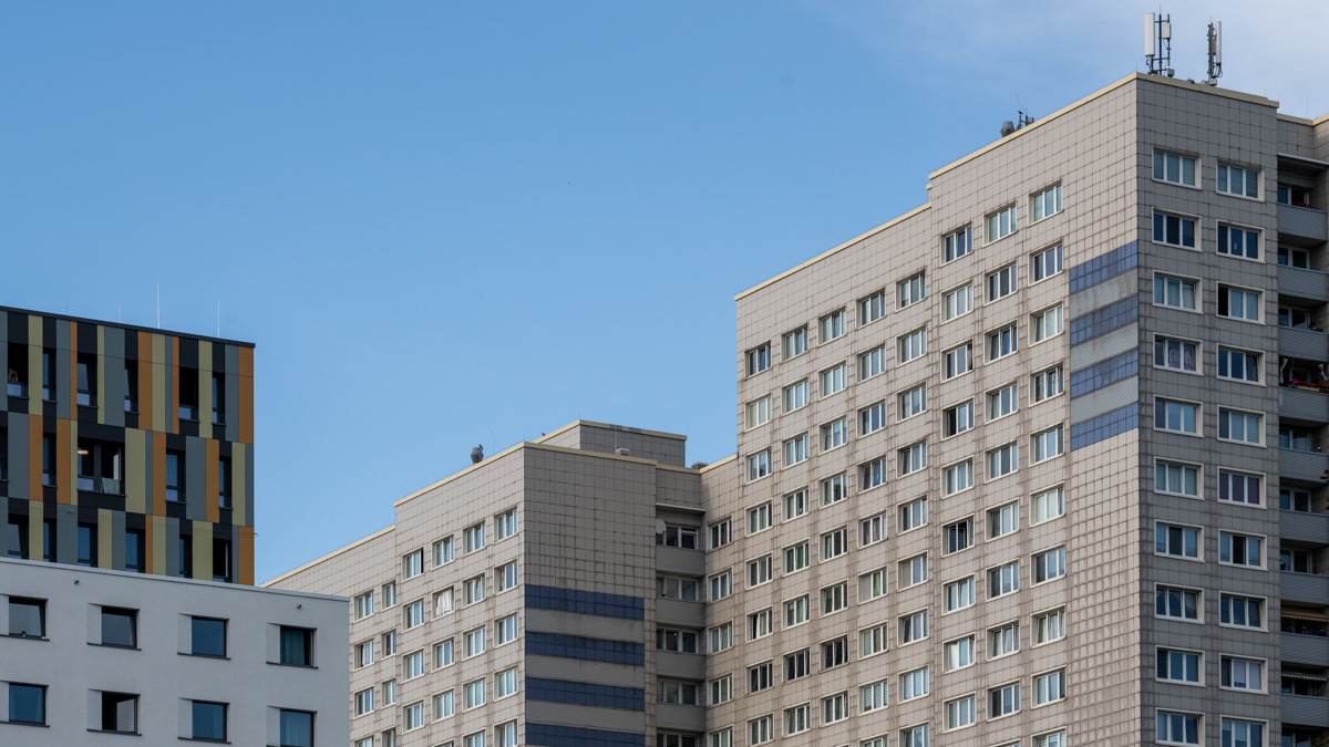 Die Aufnahme zeigt eine Reihe von modernen Wohnhäusern, die in einem geometrischen Stil gestaltet sind. Die Gebäude sind aus grauem Ziegelstein oder Beton gefertigt und mit großen Fensterflächen versehen. Der Himmel ist ein klarer, blauer Farbton. Im Vordergrund sind einige Balkone und Fenster zu erkennen. Im Hintergrund sind Antennen auf den Gebäuden zu sehen.