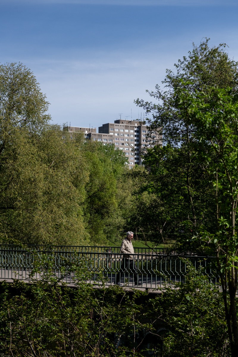 Die Aufnahme zeigt einen Mann, der auf einer Brücke in einem Park entlanggeht. Im Hintergrund erheben sich hohe, moderne Gebäude. Die Vegetation ist üppig und grün, was auf einen sonnigen Tag hindeutet. Die Szene wirkt ruhig und urban.