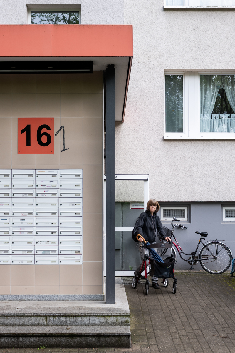 Die Aufnahme zeigt eine Frau, die in einem Rollstuhl vor einem grauen Wohnhaus sitzt. Im Vordergrund steht ein Fahrrad, und im Hintergrund sind mehrere Postboxen zu sehen. Die Gebäudefront ist in einem hellen Grau gehalten und mit einer schwarzen horizontalen Linie unterteilt. Die Szene wirkt ruhig und alltäglich.