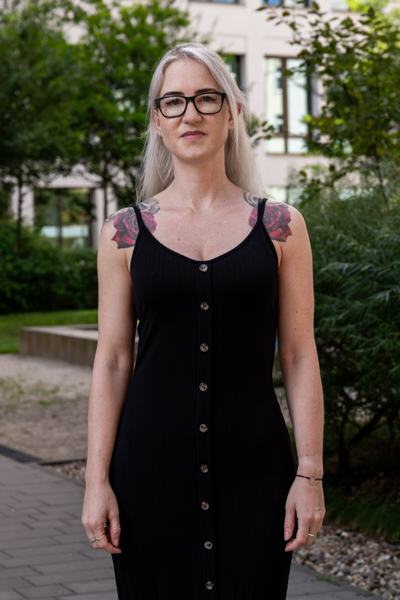 Die Aufnahme zeigt eine junge Frau mit weiß-blondem Haar, die in eine rechteckige Brille gestützt werden. Sie trägt ein schwarzes, eng anliegendes Kleid mit Trägern und einen silbernen Armreif. Ihre Gesichtszüge sind ernst und konzentriert. Im Hintergrund befindet sich ein modernes, helles Gebäude mit einer Fassade aus Putz und großen Fenstern. Die Szene ist von üppigem Grün, vermutlich Laub, umgeben. Der Boden ist mit Pflastersteinen bedeckt. Die Beleuchtung ist natürlich und diffus.