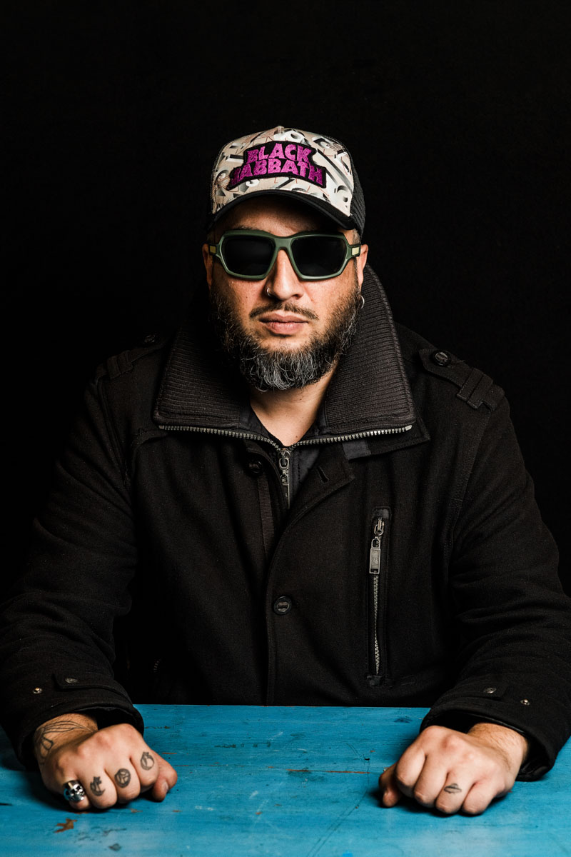 A metalhead with a short beard and with basecap and sunglasses sitting at a table.