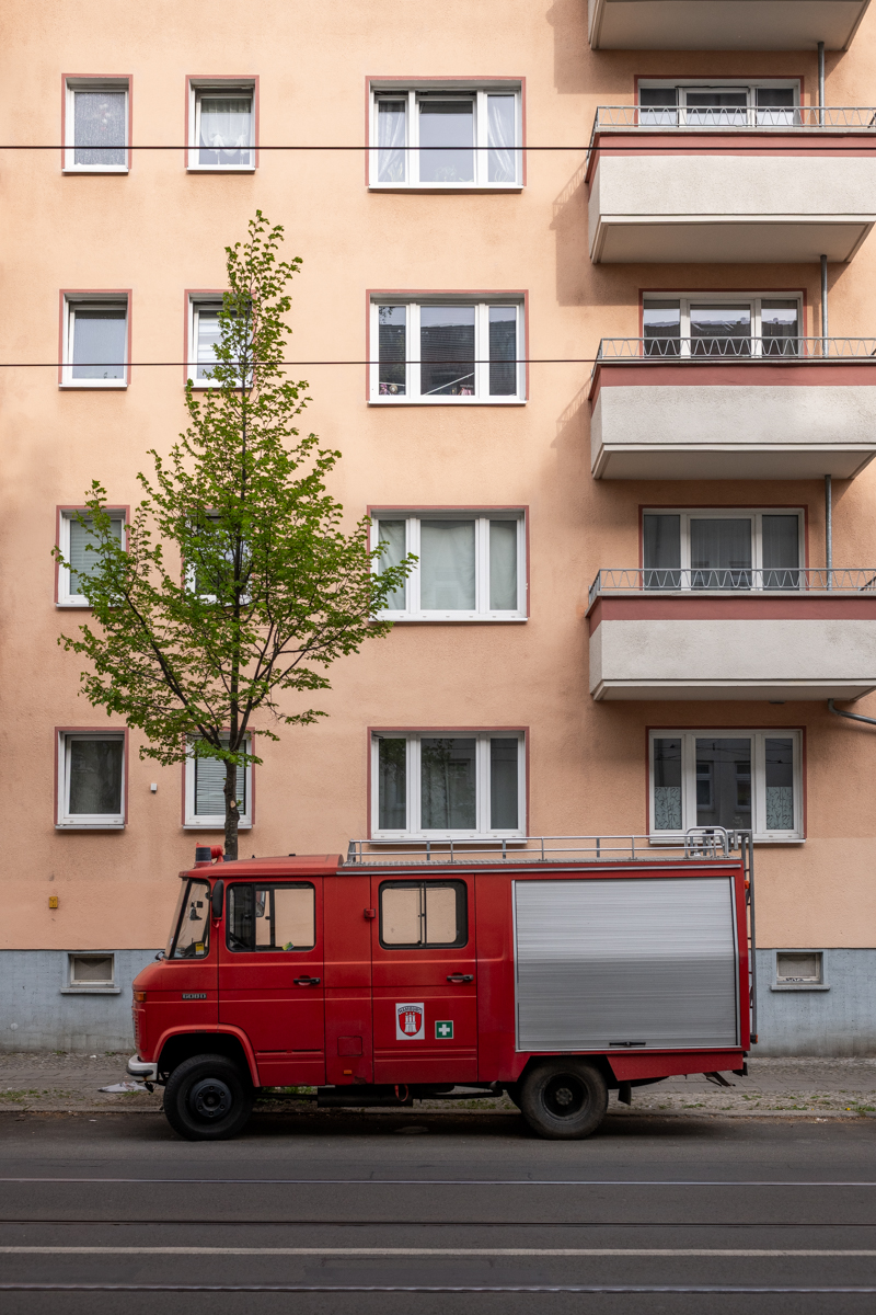 Das Bild zeigt eine straßenseitige Aufnahme eines roten Feuerwehrwagens, vermutlich ein Mercedes-Benz Unimog, der vor einem mehrgeschossigen Wohnhaus steht. Im Vordergrund steht ein Baum mit frischem, jungem Laub. Die Fassade des Wohnhauses ist in einem hellen, rosafarbenen Ton gehalten und mit vielen rechteckigen Fenstern versehen. Die Straße ist mit Stromleitungen versehen. Die Szene wirkt ruhig und urban.