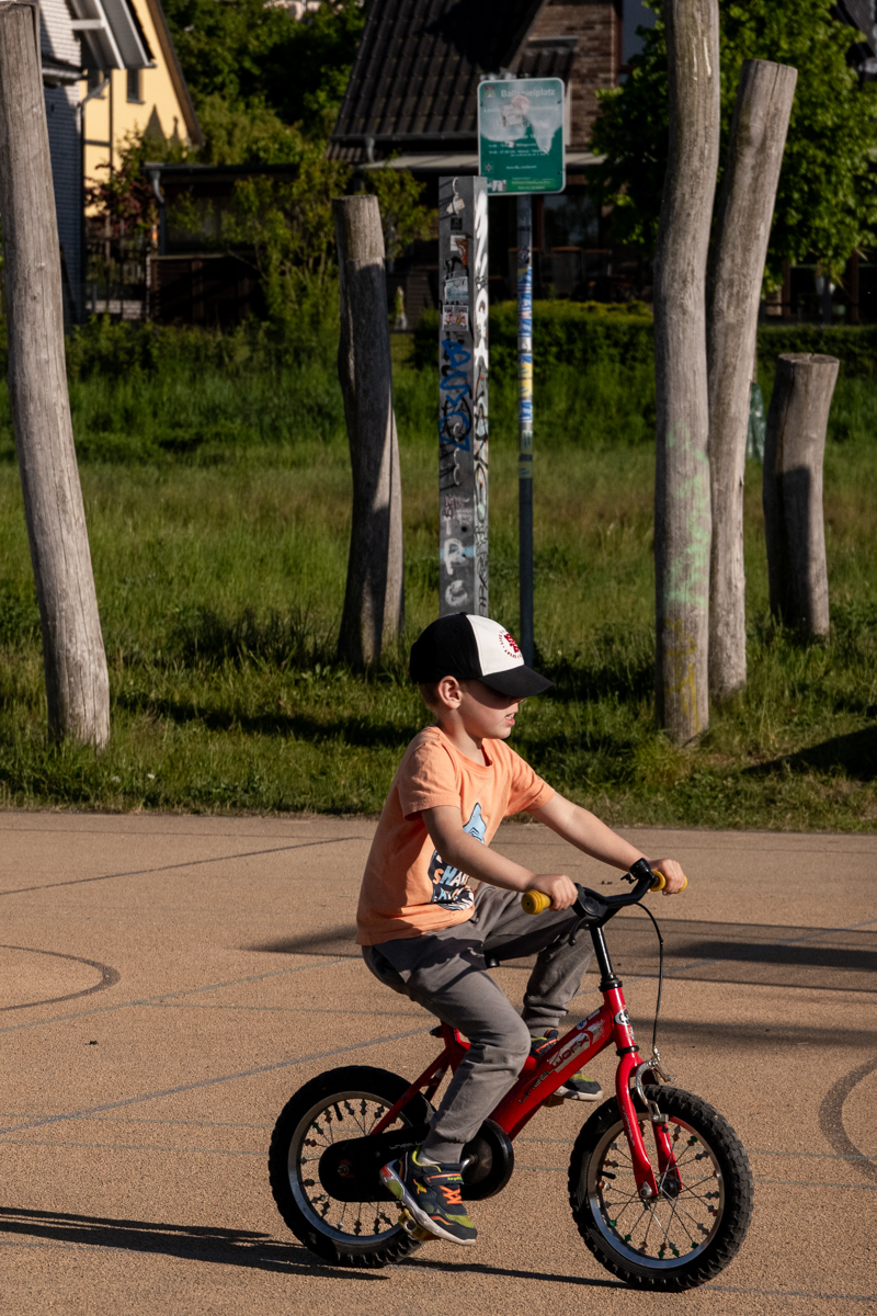 Das Bild zeigt einen jungen Mann, vermutlich im Alter von etwa 8-12 Jahren, der auf einem roten Kinderfahrrad auf einem asphaltierten Weg fährt. Er trägt einen Baseballcap, ein orangefarbenes T-Shirt mit einem Aufdruck und Jeans. Im Hintergrund befinden sich alte, verwitterte Gebäude mit Schieferdächern und Graffiti. Es sind auch Bäume und einige Holzstützen zu sehen. Der Weg ist uneben und weist Abnutzungsspuren auf. Die Lichtverhältnisse sind hell und sonnig, was auf eine warme Tageszeit hindeutet.