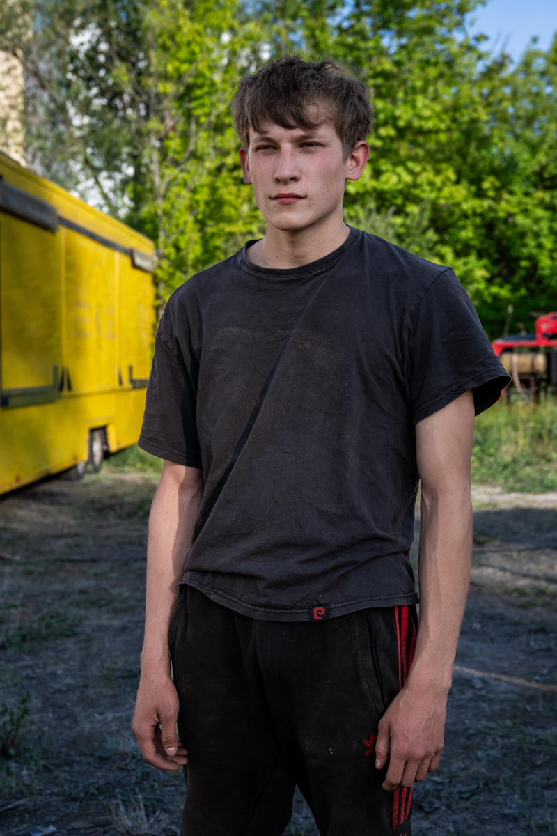 Die Aufnahme zeigt einen jungen Mann, vermutlich im Alter von etwa 20-25 Jahren, der vor einem gelben Van in einer grünen, ländlichen Umgebung steht. Er trägt ein schwarzes T-Shirt und rot-schwarze Trainingshosen. Die Szene wirkt ungestört und realistisch. Der Hintergrund ist von Bäumen und Vegetation geprägt, was auf eine ländliche oder sonstige Umgebung hindeutet.