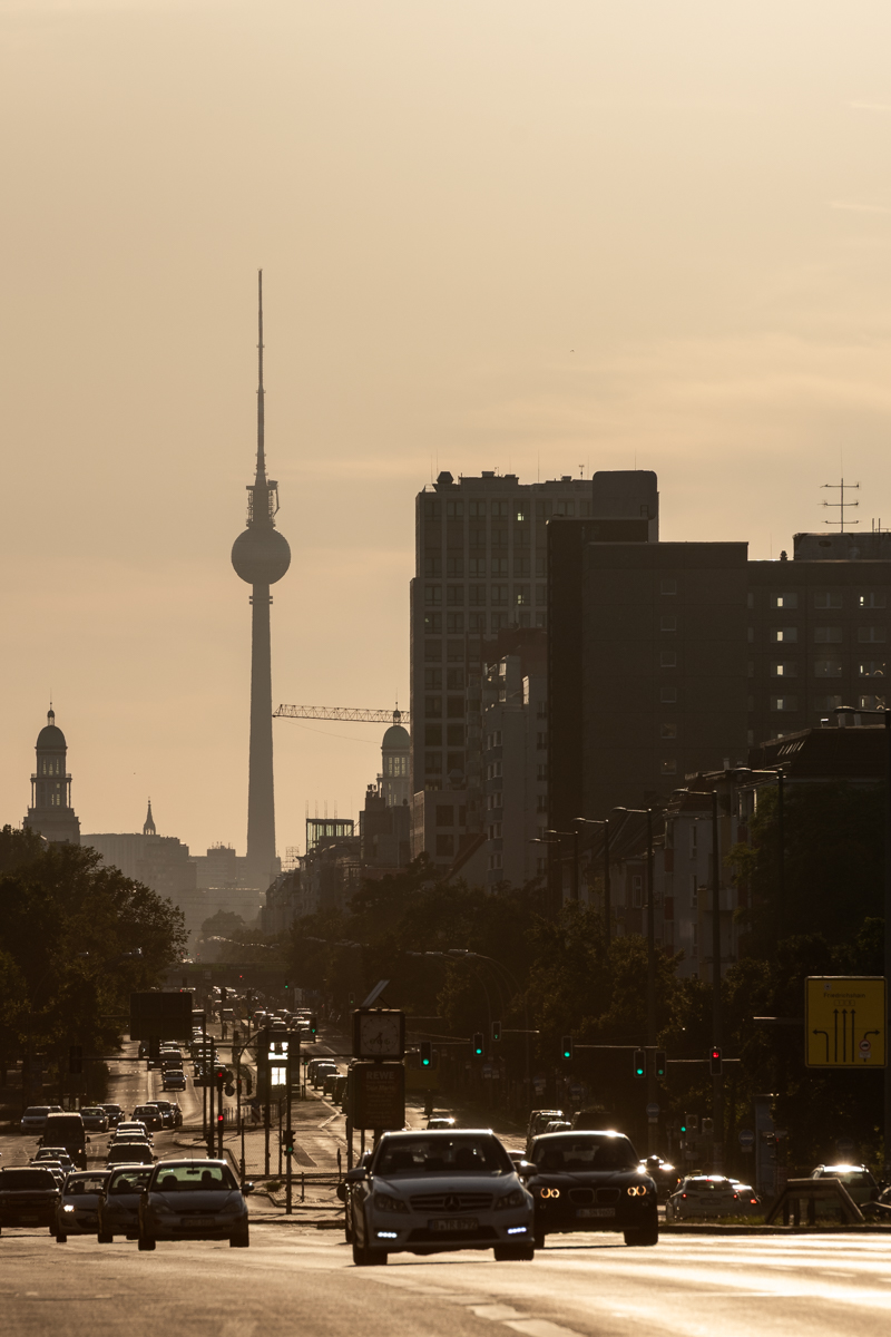 Das Bild zeigt eine belebte Straße in einer Großstadt, wahrscheinlich Berlin, bei Sonnenuntergang. Im Hintergrund sind mehrere Gebäude, darunter der Berliner Fernsehturm, silhouettiert. Die Straße ist mit zahlreichen Autos befahren, darunter Taxis. Die Stimmung ist ruhig und melancholisch, typisch für einen Sonnenuntergang.