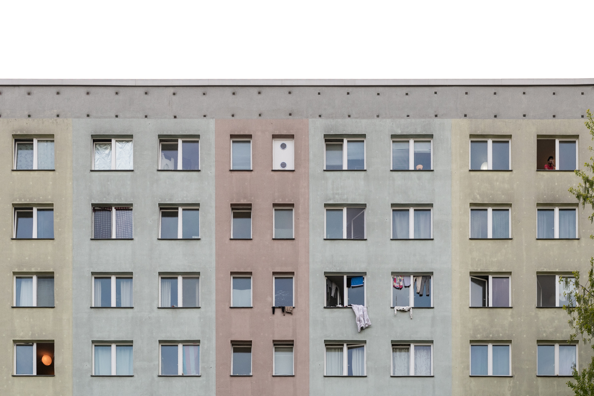 Das Bild zeigt eine mehrgeschossige Wohnhausfront in einem eher monochromen Farbton. Die Fassade ist in verschiedenen Grautönen und Rot-Braun-Tönen gehalten, was typisch für Wohngebäude der DDR-Zeit ist. Im Vordergrund sind einige Fenster mit unterschiedlich geöffneten Blenden erkennbar, die einen Eindruck von den Bewohnern vermitteln. Vor den Fenstern hängen Wäschestücke, was eine alltägliche Szene darstellt. Die Beleuchtung ist durch die Fenster unterschiedlich, was die Tiefe des Bildes betont.