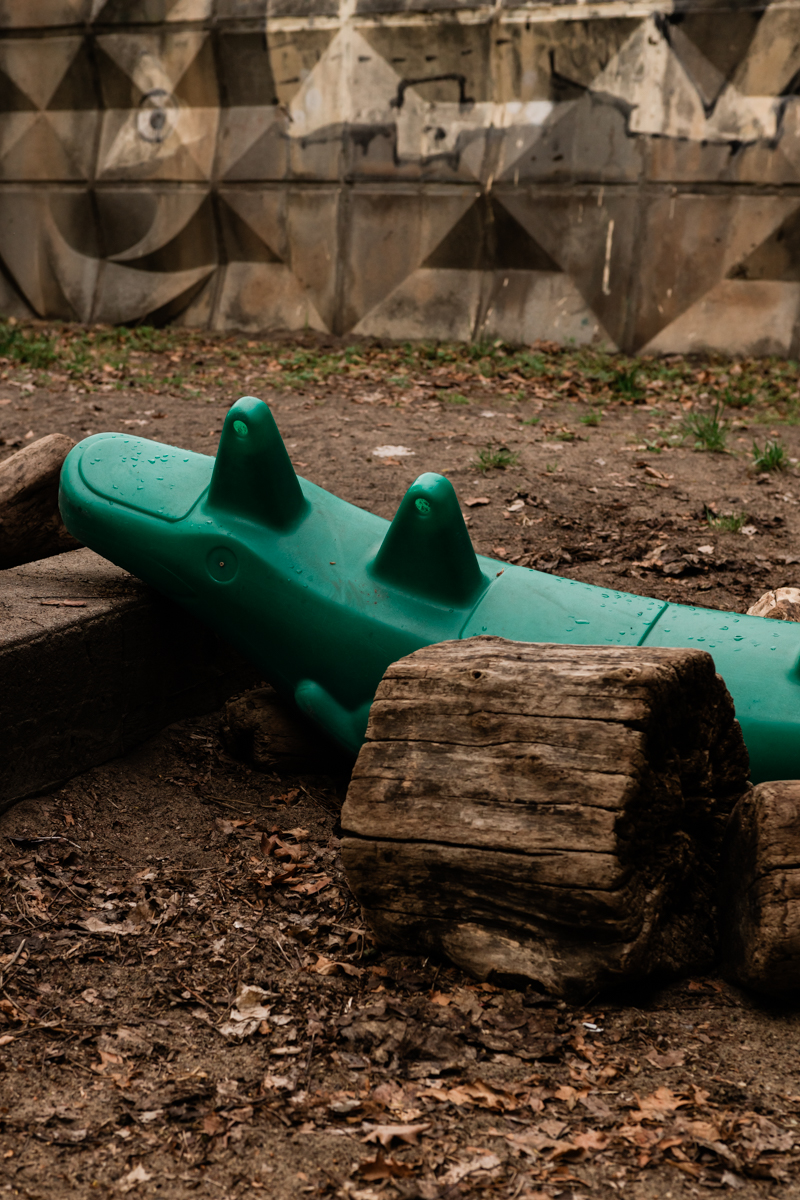 Das Bild zeigt einen grünen Schaukeldrachen, der auf einem Spielplatz in einem unebenen, mit Erde bedeckten Bereich steht. Im Hintergrund befindet sich ein Betonmauerwerk mit einem geometrischen, dreieckigen Muster. Holzklötze liegen ebenfalls auf dem Boden. Die Szene wirkt etwas düster und ungesättigt.