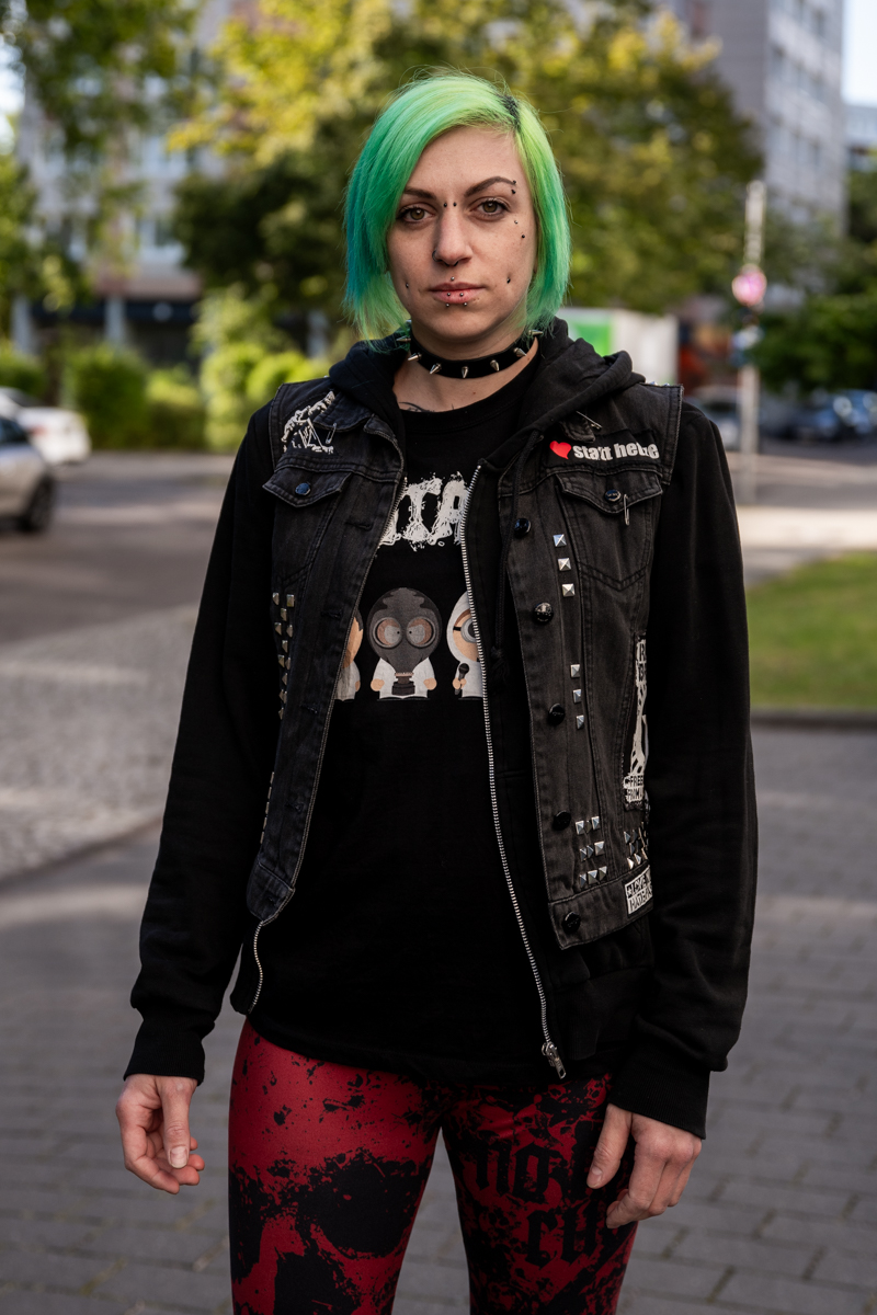 Die Aufnahme zeigt eine junge Frau mit auffälligem grünen Haar, das in einem asymmetrischen Bob geschnitten ist. Sie hat Piercings und trägt eine schwarze Denim-Jacke mit zahlreichen Patches, darunter ein Band-Logo. Darüber trägt sie einen schwarzen Hoodie mit der Aufschrift „Gutalax“. Ihre Hose ist rot und weist ein fleckiges Muster auf. Sie blickt direkt in die Kamera mit einem ernsten Gesichtsausdruck. Im Hintergrund sind moderne Gebäude zu sehen, vermutlich ein Wohngebiet. Ein graues, leicht bewölktes Tageslicht sorgt für eine neutrale Beleuchtung.