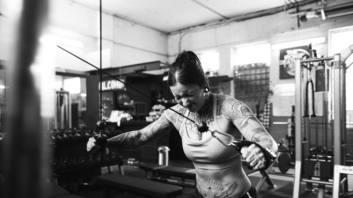 Das Schwarzweißbild zeigt eine Frau während einer Krafttrainingsübung in einem Fitnessstudio. Die Szene vermittelt Anstrengung und Konzentration.