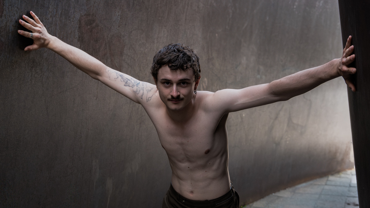 Das Bild zeigt einen Porträt-Szene eines Schauspielers, Felix Steinhilber, der vor einer grauen Metallwand mit ausgestreckten Armen steht. Die Pose vermittelt Kontemplation, Verletzlichkeit und eine gewisse Kraft. Der Hintergrund ist dunkel und unscharf, was die Aufmerksamkeit auf den Schauspieler lenkt.